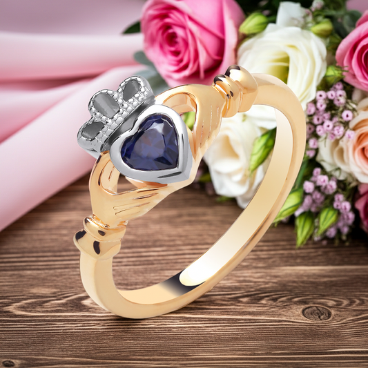 Gold Claddagh Ring with Heart-Shaped Blue Stone and Crown Design, Surrounded by Flowers